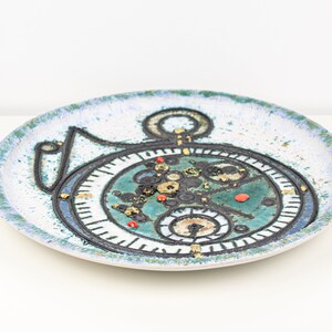 May include: A white ceramic plate with a blue and green design of a clock face with gold accents.