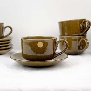 Könnte beinhalten: Satz mit sechs braunen Keramik-Teetassen und Untertassen mit goldenen Akzenten. Die Tassen haben eine abgerundete Form und einen Henkel an der Seite. Die Untertassen sind leicht gewölbt und haben ein passendes Design.