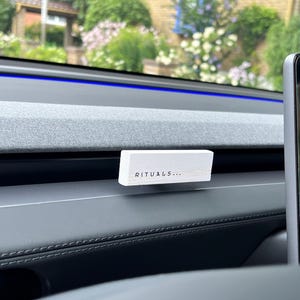 Puede incluir: Un clip ambientador de coche rectangular de color blanco con la palabra RITUALS... impresa en negro en la parte frontal. El objeto está sujeto a la rejilla de ventilación horizontal del salpicadero de un vehículo gris oscuro, junto a una pantalla.