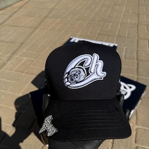 May include: Black baseball cap with a flat brim. The cap features a white embroidered design of the letters "Ch" with a skull. A silver pin with the text "Solstice" is on the side. The cap is held by a gloved hand.