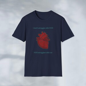 Könnte beinhalten: Marineblaues T-Shirt mit roter anatomischer Herzgrafik. Der Text "I don't struggle with POTS" steht über dem Herzen und "POTS struggles with me" darunter. Der Text und das Design sind türkis.