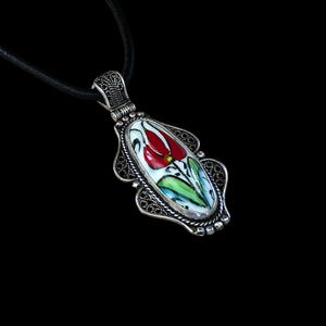 May include: A silver pendant necklace with a black leather cord. The pendant features a white oval with a red and green floral design.