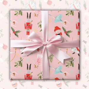 May include: Pink wrapping paper with a repeating pattern of nutcracker soldiers, ballerinas, holly, and snowflakes. A pink ribbon is tied around the paper with a bow in the center.