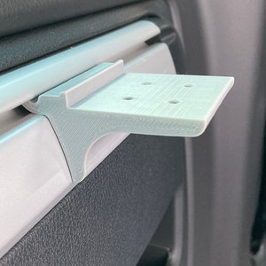 Puede incluir: Un soporte para teléfono gris impreso en 3D diseñado para encajar en la ventilación de un coche.