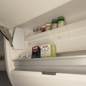 Könnte beinhalten: Weißer Küchenschrank mit einem herunterklappbaren Regal. Das Regal ist gefüllt mit verschiedenen Gläsern und Schachteln mit Lebensmitteln, darunter Gewürzgläser, Teebeutel und eine Schachtel "HiPP Bio Fenchel Tee".