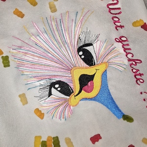 May include: A white fabric tote bag featuring a colourful embroidered design of a cartoon bird's face. The bird has large eyes, a yellow beak, and a blue head. The text "Wat guckst du?" is embroidered in black on the bag. The bag is surrounded by scattered gummy bears in various colours.