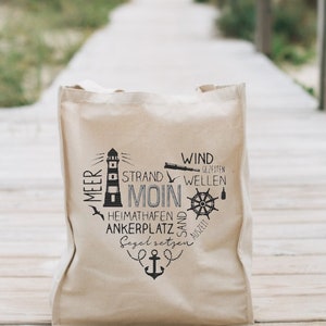 Op de afbeelding: Een beige canvas draagtas met een zwart grafisch ontwerp met een vuurtoren, nautische symbolen en de tekst "Strand Moin" en andere Duitse woorden.