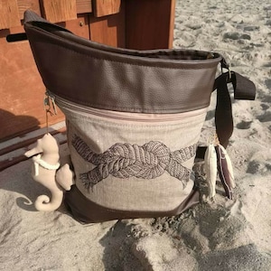 Op de afbeelding: Een bruine leren en beige canvas draagtas met een ritssluiting en een lange bruine band. De tas heeft een geborduurd ontwerp van een touwknoop. De tas ligt op een zandstrand.