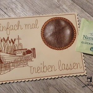 May include: A beige coaster with brown embroidery depicting a boat, reeds, and the text "Einfach mal treiben lassen". The coaster has a brown leather circle in the center. A green tea bag with the text "NamasTee Ayurvedische Mischung Glücksgefühle" is visible in the background.