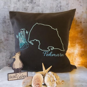 Puede incluir: Una almohada marrón oscuro con un diseño bordado de la isla de Fehmarn, Alemania, con un barco y gaviotas. La almohada está sobre una superficie blanca con conchas y una estrella de mar. El texto "Insel Leben" está en un pequeño letrero de madera.