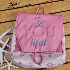Könnte beinhalten: Ein rosa Rucksack mit einem rosa-weißen gemusterten Boden. Die vordere Klappe ist mit den Worten "Be You tiful" in grauer Farbe bestickt.