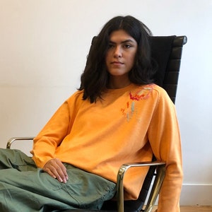 May include: An orange long-sleeve sweater with floral embroidery on the shoulder, paired with olive green cargo pants. The person is seated in a black leather chair with a silver base, resting on a patterned rug.