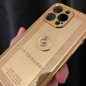May include: A gold-colored phone case with a decorative lion emblem and the text "Joy Kleimann." The case has a geometric border design and the text "24K GOLD EXCULUSIVE EDITION 1973 ETC. ARR INC."