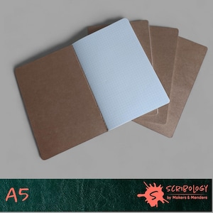 May include: An A5 dot grid notebook with a brown cover and light blue interior pages. The notebook is open, revealing the dotted pages, with additional notebooks visible behind it. The text "A5" is in the lower left corner.