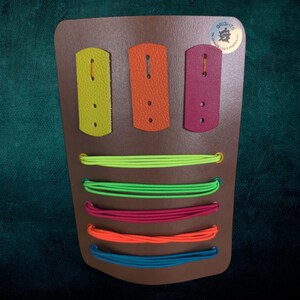May include: A brown leather board with three colourful leather straps and five colourful elastic bands. The board has the text "Brights" and a logo with the text "The crafting & workshop".