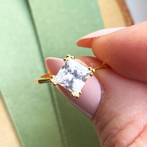 Peut inclure: Une bague en or avec une pierre précieuse carrée et transparente. La bague a un fin anneau et de petits éléments décoratifs autour de la pierre précieuse. La bague est tenue sur un fond vert.