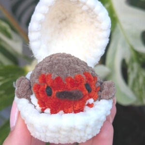 May include: A crocheted creature in a white shell. The creature has a brown head, a red face, and black eyes. The shell is open, showing the creature. The background is a blurred green plant.