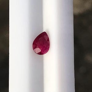 Può includere: Una gemma di rubino sfaccettata a forma di pera con un colore rosso intenso. La pietra presenta una parte superiore appuntita e una base arrotondata, mostrando molteplici sfaccettature geometriche che riflettono la luce sulla sua superficie, posizionata su una superficie bianca.