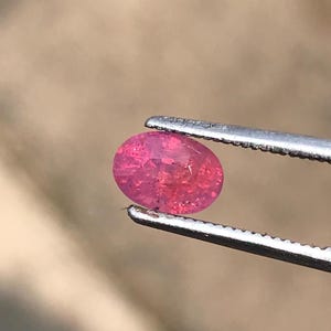 May include: A pink oval-shaped gemstone held by tweezers. The gemstone has a slightly cloudy appearance.