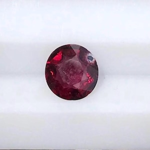 May include: A round, faceted gemstone with a deep, saturated red color. The stone features a brilliant cut with multiple triangular and kite-shaped facets that reflect light, creating a complex pattern of dark and bright crimson tones throughout.