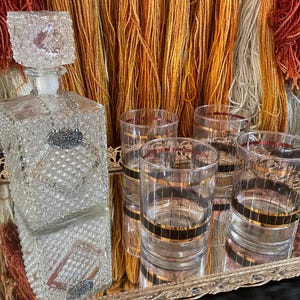 May include: A set of four clear glass tumblers with gold and black stripes, and a clear glass decanter with a diamond pattern and a silver tag that says "Scotch". The glasses and decanter are on a gold metal tray.