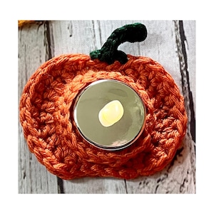 May include: A crocheted pumpkin-shaped candle holder with a round, silver-coloured lid. The lid has a small, white, round wax melt inside.