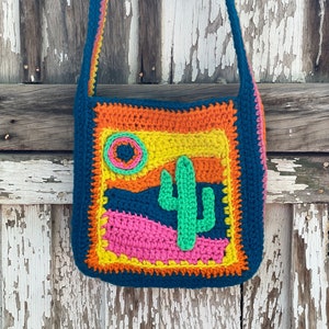 Puede incluir: Un bolso de hombro de crochet con un diseño de escena del desierto colorido. El bolso está hecho con hilo azul, naranja, amarillo, verde, rosa y blanco. El diseño presenta un cactus, un sol y una cordillera.