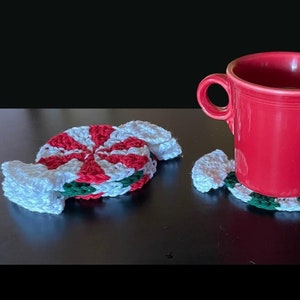 May include: Two crocheted peppermint candy coasters in red, white and green. A red mug is sitting on one of the coasters.