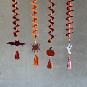 May include: Four crocheted Halloween decorations hanging from the ceiling. Each decoration features a different Halloween theme: a bat, a spider, a pumpkin, and a ghost. The decorations are made with orange, black, white, and purple yarn.