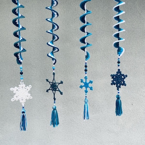 Könnte beinhalten: Vier handgefertigte gehäkelte Spiralornamente in Blau- und Weißtönen, jeweils mit einem Schneeflocken- und Quastenanhänger. Die Ornamente sind vor einem grauen Hintergrund aufgehängt. Die Schneeflocken und Quasten verleihen dem Spiraldesign eine dekorative Note.