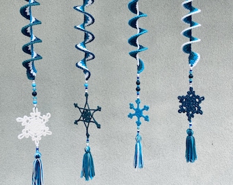 Könnte beinhalten: Vier handgefertigte gehäkelte Spiralornamente in Blau- und Weißtönen, jeweils mit einem Schneeflocken- und Quastenanhänger. Die Ornamente sind vor einem grauen Hintergrund aufgehängt. Die Schneeflocken und Quasten verleihen dem Spiraldesign eine dekorative Note.