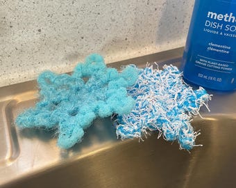 Puede incluir: Dos estropajos de fregar azules y blancos con forma de estrella. Los estropajos están sobre un fregadero de acero inoxidable. Una botella de jabón para lavar platos está en el fondo.