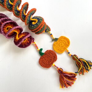 Puede incluir: Dos guirnaldas de calabaza de ganchillo con hilo naranja, amarillo, verde y morado. Cada guirnalda tiene una calabaza de ganchillo con un borla de cuentas.