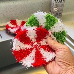 May include: Circular, handmade dish scrubbers in red, white, and green. The scrubbers have a textured, loopy appearance. One is held in a hand, while another rests on a metal surface. A bottle of dish soap is in the background.