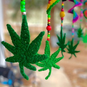 May include: A green crocheted marijuana leaf hanging from a beaded string with colorful beads. The leaf is a popular symbol of the cannabis culture.