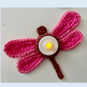 Puede incluir: Una libélula de crochet rosa y marrón con un portavelas de té blanco en el centro. La vela de té está encendida y brillando.