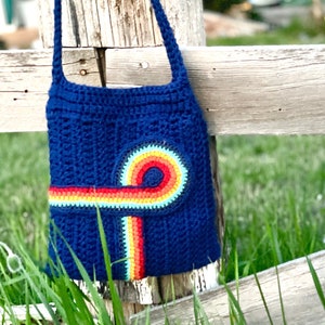 Può includere: Una borsa di uncinetto blu con un design arcobaleno. La borsa ha una lunga tracolla ed è appesa a una recinzione di legno.
