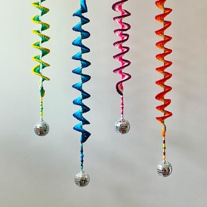 May include: Four colorful, spiral-shaped crochet decorations hanging from strings. Each spiral is a different color: yellow and green, blue, pink and purple, and orange and red. A small disco ball hangs at the bottom of each spiral.