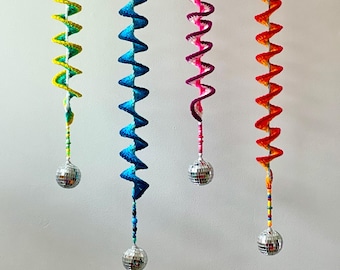 Può includere: Quattro decorazioni all'uncinetto a forma di spirale colorata appese a fili. Ogni spirale è di un colore diverso: giallo e verde, blu, rosa e viola, e arancione e rosso. Una piccola palla da discoteca è appesa in fondo a ogni spirale.