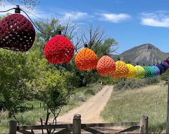 May include: A string of colorful, crocheted light covers in a rainbow of hues, suspended outdoors. The lights are strung across a dirt road, with a mountain in the background. The colors include burgundy, red, orange, yellow, green, blue, and purple.
