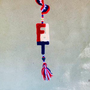 May include: A handmade crochet wall hanging featuring the letter "F" in red, white, and blue. The letter is connected to a tassel with red, white, and blue yarn and beads. The top of the hanging has a decorative element with the same colors.