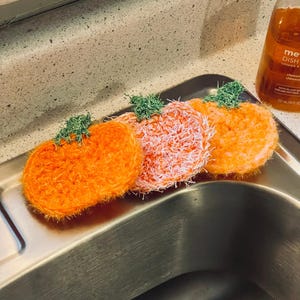 Peut inclure: Trois éponges en forme de citrouille orange avec des tiges vertes sont posées dans un évier en acier inoxydable. Les éponges sont faites d'un matériau texturé et fibreux. Une bouteille de liquide vaisselle est visible en arrière-plan.
