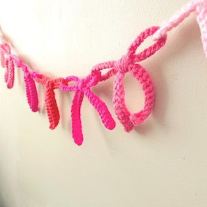 May include: A handmade crochet garland featuring a series of pink bows in varying shades. The bows are connected by a crocheted chain, creating a decorative banner. The color palette includes light, medium, and bright pink hues.
