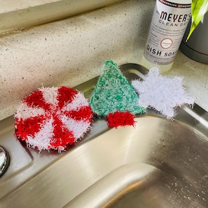 Op de afbeelding: Een verzameling handgemaakte gehaakte scrubbies in feestelijke vormen. Inclusief een rood-witte pepermuntwervel, een groene kerstboom met een rode basis en een witte sneeuwvlok. Een fles Meyer's Clean Day afwasmiddel staat op de achtergrond.
