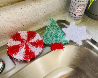 Puede incluir: Colección de estropajos de ganchillo hechos a mano con formas festivas. Incluye un remolino de menta roja y blanca, un árbol de Navidad verde con base roja y un copo de nieve blanco. Una botella de jabón para platos Meyer's Clean Day está en el fondo.
