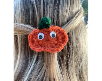 Può includere: Zucca arancione all'uncinetto con occhietti e un gambo verde. La zucca è attaccata a un elastico per capelli.