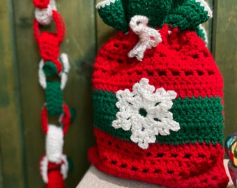 May include: A red and green crocheted Christmas stocking with a white snowflake design. The stocking is hanging on a wooden wall with a red and white crocheted garland.