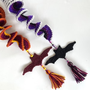 May include: Three crocheted bat ornaments with colourful yarn tails. The bats are purple, black, and maroon. The tails are made of yarn in shades of orange, yellow, and white.