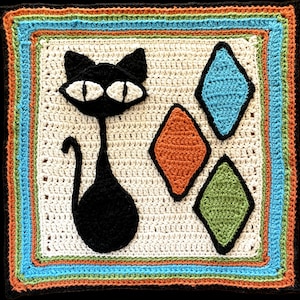 Op de afbeelding: Een wit gehaakt vierkant met een zwarte kat, oranje, blauwe en groene ruitvormen. De kat heeft grote ogen en een lange staart.
