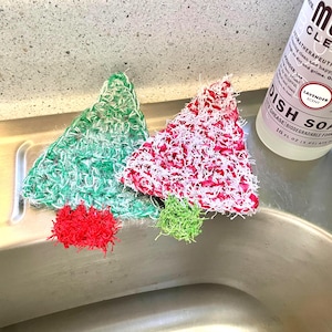 Op de afbeelding: Twee handgemaakte, kerstboomvormige scrubbies in groen en rood met witte accenten, en twee kleinere scrubbies in rood en groen. Een fles lavendelgeurende afwasmiddel is zichtbaar op de achtergrond.